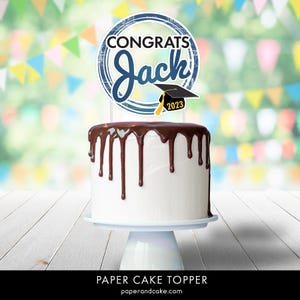 May include: A blue and white paper cake topper with the text "Congrats Jack" and a graduation cap with the year 2023. The topper is on top of a white cake with chocolate frosting.