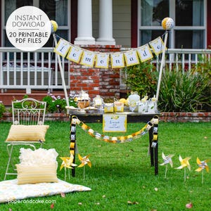 May include: A lemonade stand set up in a backyard with a black table, yellow and white striped paper chains, and a banner that reads "Lemonade". There are yellow and white pinwheels on the ground and a white chair with a yellow and white striped pillow.