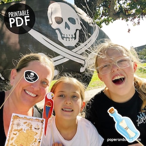 May include: Three people are smiling for a photo in front of a pirate flag. The flag is black with a white skull and crossbones. The people are holding pirate-themed props, including a treasure map, a parrot, and a bottle with a message.