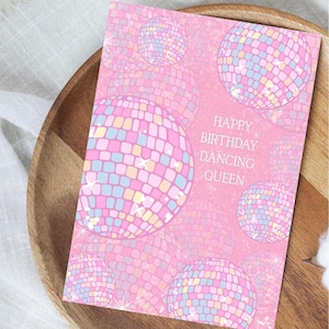 May include: Pink birthday card with the text "Happy Birthday Dancing Queen" and a pattern of colorful disco balls.