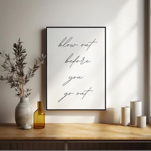 Könnte beinhalten: Schwarz-weißes Typografie-Druckbild mit dem Text "blow out before you go out". Der Druck ist in einem schwarzen Rahmen gerahmt und hängt an einer weißen Wand.