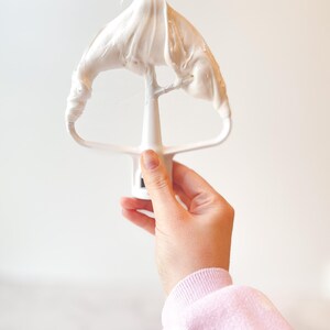 May include: A white mixer attachment covered in white meringue. The attachment is held by a hand wearing a pink sweatshirt. The background is a plain white surface.