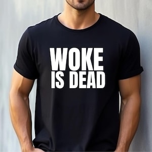May include: Black t-shirt featuring the white text "WOKE IS DEAD". The text is in a bold, sans-serif font. The shirt has short sleeves and a crew neck. The image shows the shirt on a person.