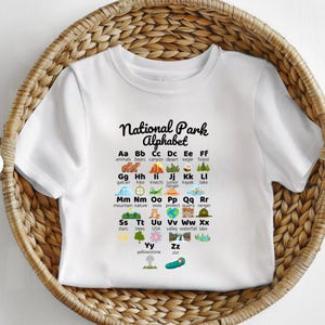 May include: White t-shirt featuring the text "National Park Alphabet" with an illustrated alphabet. Each letter is paired with a park-related word and image, such as "bears" and "kayak". The shirt is displayed in a woven basket.