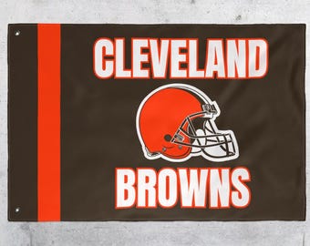 May include: Brown flag with the words "CLEVELAND BROWNS" in white block letters. The flag features a brown football helmet with an orange shell and white facemask. A vertical orange stripe is on the left side.