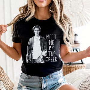 May include: Black t-shirt with a black and white image of a man and the text "MEET ME BY THE CREEK" in white. The man is wearing a plaid shirt over a white t-shirt.