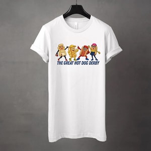 May include: White t-shirt featuring a cartoon graphic of four anthropomorphic hot dogs in various outfits. The text "THE GREAT HOT DOG DERBY" is printed below the characters in blue. The shirt has short sleeves and a crew neck.