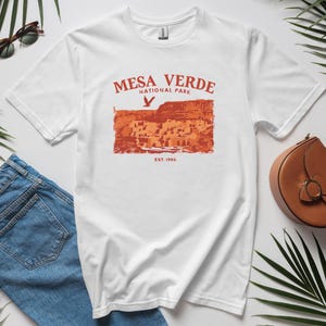 May include: White t-shirt with a red graphic print of Mesa Verde National Park, featuring a cliff dwelling and a bird flying overhead. The text "Mesa Verde National Park" and "Est. 1906" are printed below the image.