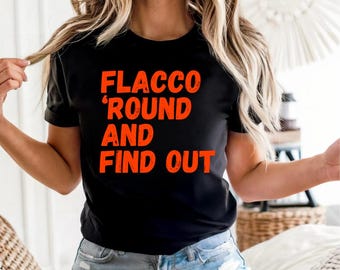 May include: Black t-shirt with the orange text "FLACCO 'ROUND AND FIND OUT". The shirt has short sleeves and a crew neck. The shirt is worn with denim shorts.