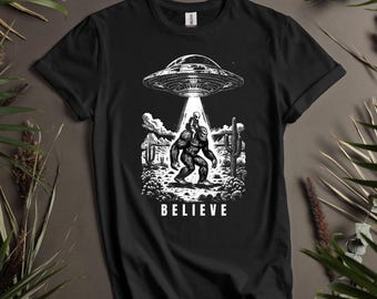May include: Black t-shirt with a white graphic of a Bigfoot carrying an alien on its back. The graphic also includes a UFO, cacti, and the word "BELIEVE".