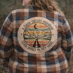 May include: A brown, blue, and orange plaid flannel shirt with a circular graphic on the back. The graphic features a desert landscape with a road, cactus, and the text "A Dark Desert Highway, Cool Wind In My Hair."