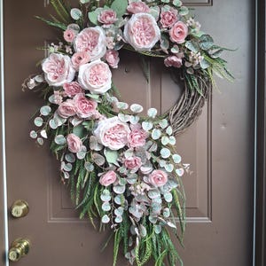 May include: A decorative wreath featuring a circular grapevine base adorned with large, light pink roses and smaller pink buds. Greenery includes eucalyptus and fern-like leaves, creating a full, natural look. The wreath is designed for door or wall decor.