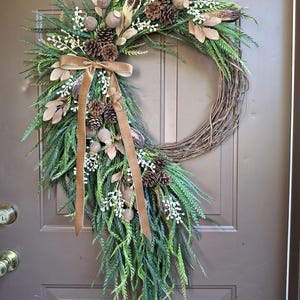 May include: A decorative wreath with a brown grapevine base, adorned with green faux foliage, pinecones, and tan accents. A brown velvet ribbon is tied in a bow. The wreath is displayed on a brown door.