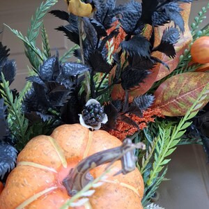 May include: A decorative arrangement featuring a bright orange pumpkin with a brown stem, surrounded by dark black foliage, textured black berry clusters, green fern-like sprigs, and autumn-toned leaves in shades of orange, red, and brown.
