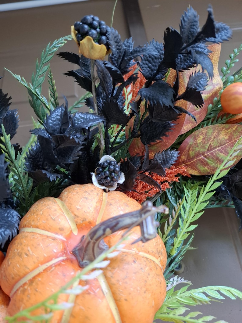 May include: A decorative arrangement featuring a bright orange pumpkin with a brown stem, surrounded by dark black foliage, textured black berry clusters, green fern-like sprigs, and autumn-toned leaves in shades of orange, red, and brown.