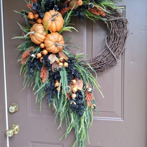 May include: A decorative wreath featuring a natural brown grapevine base adorned with three orange pumpkins, black foliage, green fern-like sprigs, rust-colored grasses, and small orange berries. The arrangement is asymmetrical and hangs vertically.
