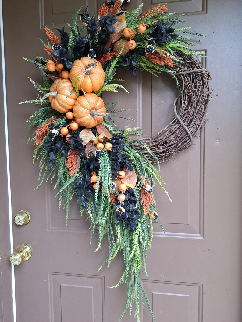 May include: A decorative wreath featuring a natural brown grapevine base adorned with three orange pumpkins, black foliage, green fern-like sprigs, rust-colored grasses, and small orange berries. The arrangement is asymmetrical and hangs vertically.
