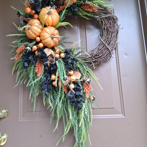 May include: A decorative wreath featuring a natural brown twig base adorned with three orange pumpkins, black foliage, green fern-like sprigs, and small orange berries. The arrangement is clustered along one side, creating a cascading effect.