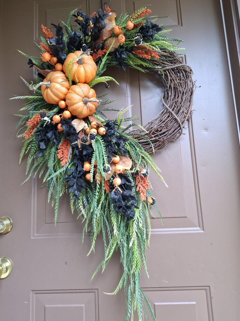 May include: A decorative wreath featuring a natural brown twig base adorned with three orange pumpkins, black foliage, green fern-like sprigs, and small orange berries. The arrangement is clustered along one side, creating a cascading effect.