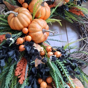 May include: A decorative wreath featuring three orange pumpkins, clusters of small orange berries, and dark black foliage. The arrangement is set on a natural brown twig base and includes textured green sprigs and small black berry accents.