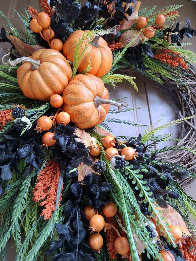 May include: A decorative wreath featuring three orange pumpkins, clusters of small orange berries, and dark black foliage. The arrangement is set on a natural brown twig base and includes textured green sprigs and small black berry accents.