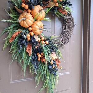 May include: A decorative wreath featuring three orange pumpkins, black foliage, green fern-like leaves, and orange berry clusters arranged on a circular brown twig base. The arrangement is asymmetrical, with the pumpkins clustered on the upper left side.