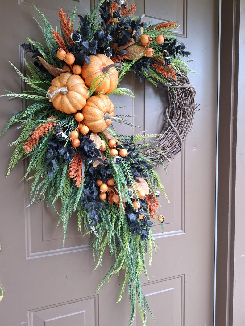 May include: A decorative wreath featuring three orange pumpkins, black foliage, green fern-like leaves, and orange berry clusters arranged on a circular brown twig base. The arrangement is asymmetrical, with the pumpkins clustered on the upper left side.