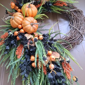 May include: A decorative wreath featuring a natural brown grapevine base adorned with three orange pumpkins, black foliage, green fern-like sprigs, orange textured floral accents, and small dark berries arranged in a cascading asymmetrical design.