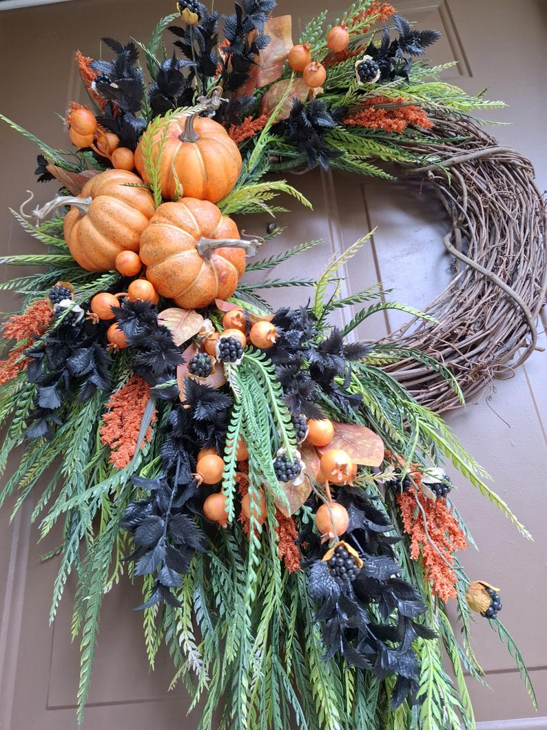 May include: A decorative wreath featuring a natural brown grapevine base adorned with three orange pumpkins, black foliage, green fern-like sprigs, orange textured floral accents, and small dark berries arranged in a cascading asymmetrical design.