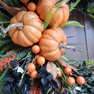 May include: A decorative wreath featuring three orange pumpkins, clusters of small orange berries, dark black foliage, green fern-like sprigs, and small dark berry bunches. The arrangement is dense with contrasting textures and colors.