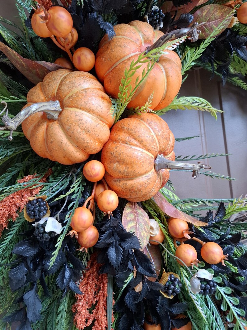 May include: A decorative wreath featuring three orange pumpkins, clusters of small orange berries, dark black foliage, green fern-like sprigs, and small dark berry bunches. The arrangement is dense with contrasting textures and colors.