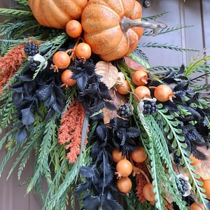 May include: A decorative wreath featuring orange pumpkins, small orange berries, and clusters of black berries. The arrangement includes green fern-like foliage, textured black leaves, and orange feathery plumes, creating a dense, layered botanical display.