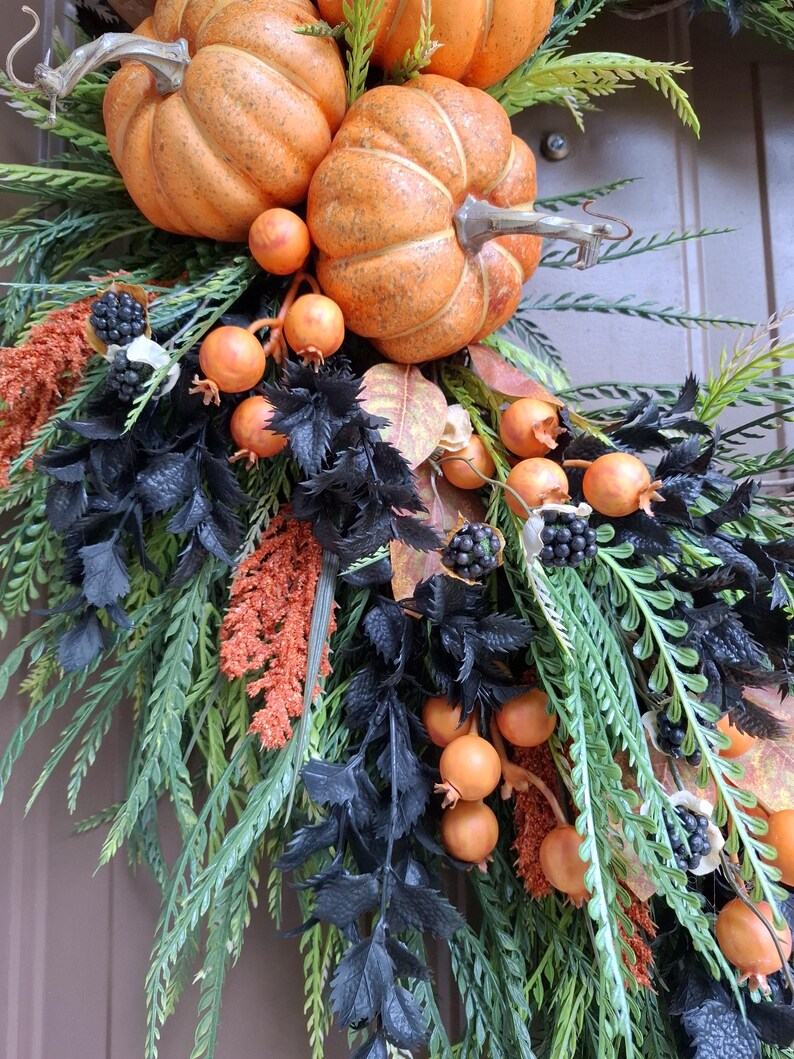 May include: A decorative wreath featuring orange pumpkins, small orange berries, and clusters of black berries. The arrangement includes green fern-like foliage, textured black leaves, and orange feathery plumes, creating a dense, layered botanical display.