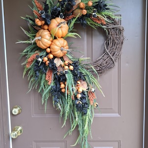 May include: A decorative wreath featuring a natural grapevine base adorned with three orange pumpkins, black foliage, green fern-like sprigs, and clusters of small orange berries. The arrangement is asymmetrical, cascading downwards along the side.