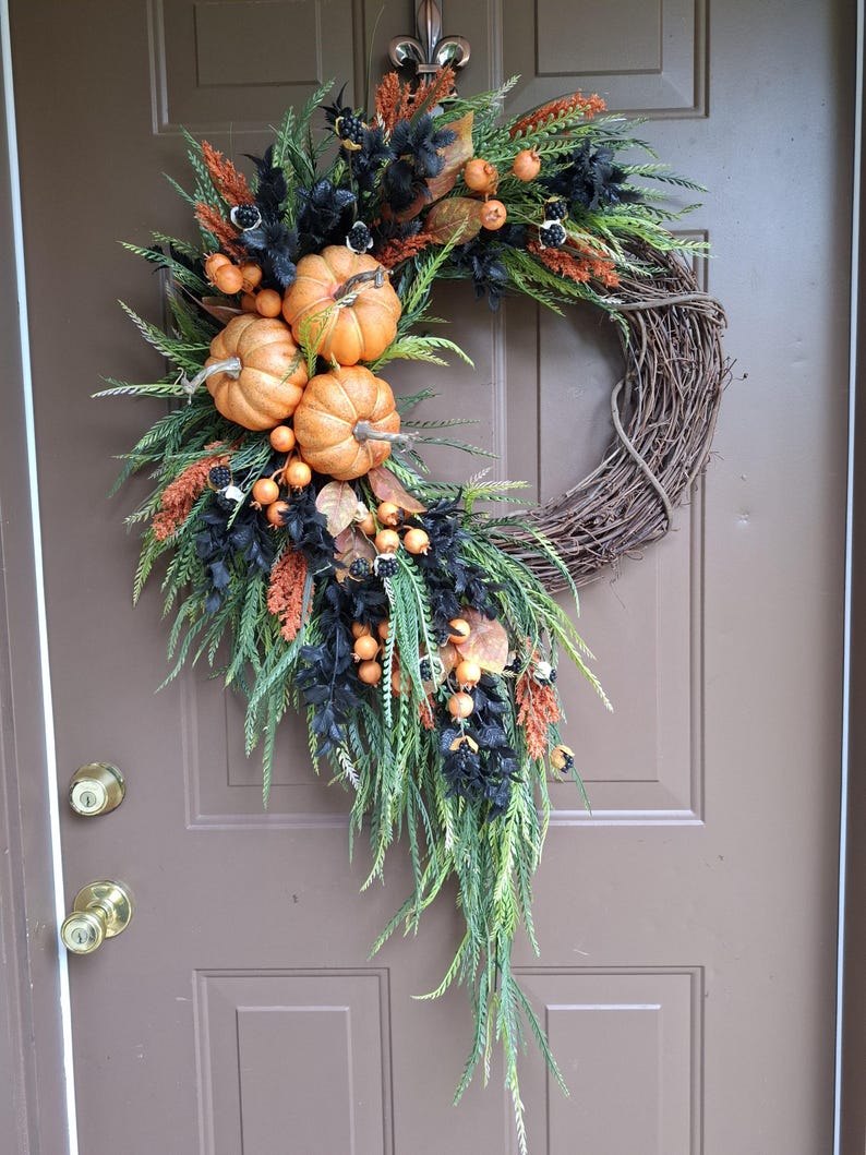 May include: A decorative wreath featuring a natural grapevine base adorned with three orange pumpkins, black foliage, green fern-like sprigs, and clusters of small orange berries. The arrangement is asymmetrical, cascading downwards along the side.