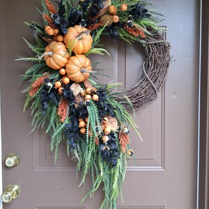 May include: A decorative wreath featuring a natural twig base adorned with artificial orange pumpkins, black foliage, green fern-like sprigs, and small orange berries. The arrangement is asymmetrical, cascading down one side of the circular frame.