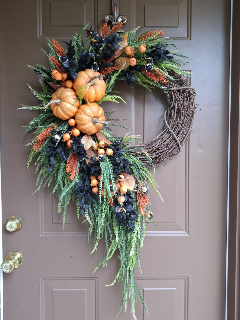 May include: A decorative wreath featuring a natural twig base adorned with artificial orange pumpkins, black foliage, green fern-like sprigs, and small orange berries. The arrangement is asymmetrical, cascading down one side of the circular frame.