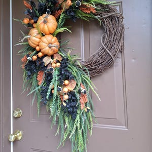May include: A decorative wreath featuring a natural brown grapevine base adorned with three orange pumpkins, black foliage, green fern-like sprigs, orange berry clusters, and small black berries arranged in a cascading asymmetrical design.
