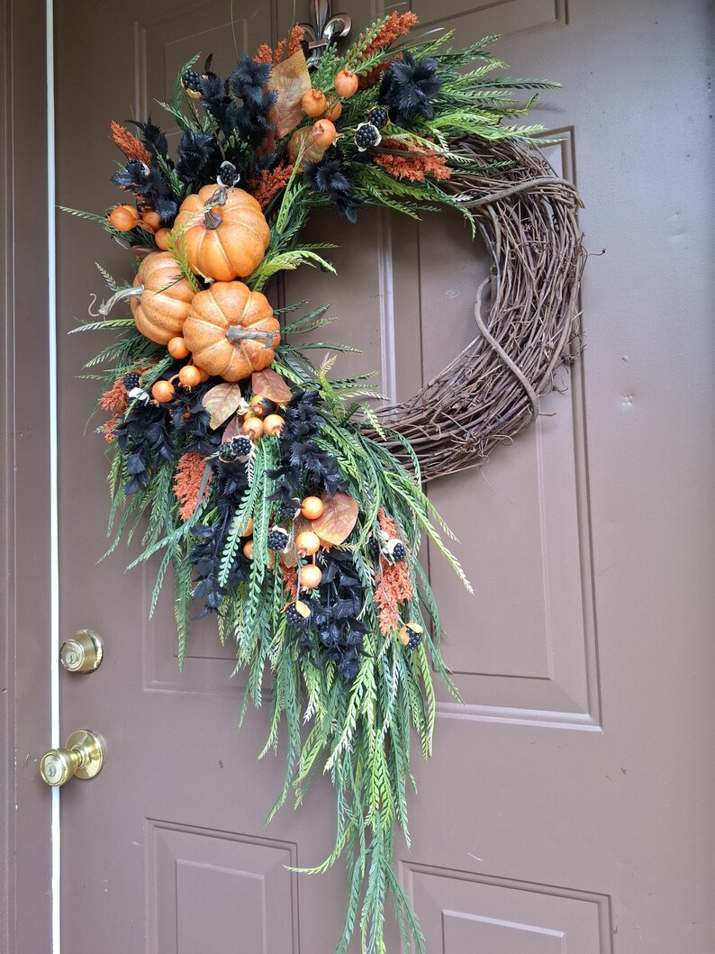 May include: A decorative wreath featuring a natural brown grapevine base adorned with three orange pumpkins, black foliage, green fern-like sprigs, orange berry clusters, and small black berries arranged in a cascading asymmetrical design.