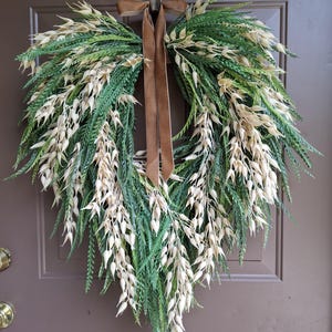 May include: A heart-shaped wreath constructed from green fern-like foliage and cream-colored oat-like stalks. A brown velvet ribbon is tied at the top center, with two long tails hanging down through the middle of the heart-shaped arrangement.