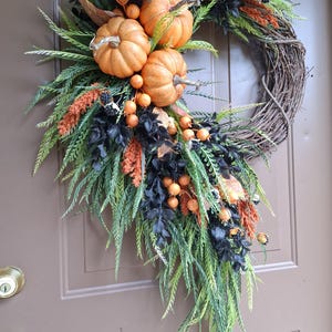 May include: A decorative wreath featuring a natural grapevine base adorned with three orange pumpkins, black foliage, green fern-like sprigs, orange berry clusters, and small black berries, arranged in an asymmetrical cascading design.