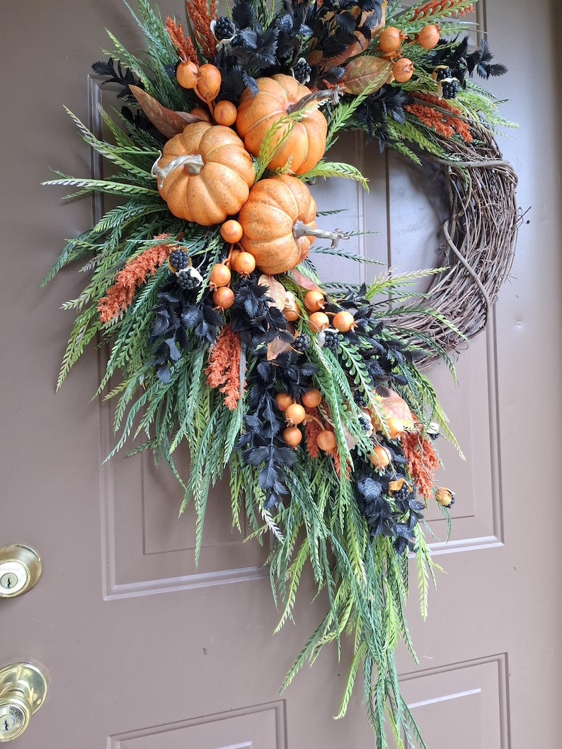 May include: A decorative wreath featuring a natural grapevine base adorned with three orange pumpkins, black foliage, green fern-like sprigs, orange berry clusters, and small black berries, arranged in an asymmetrical cascading design.