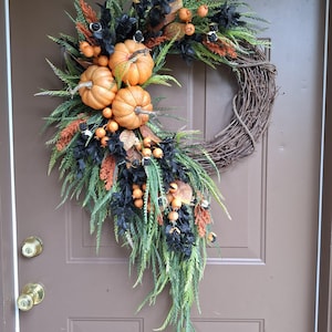 May include: A decorative wreath featuring a circular brown twig base adorned with three orange pumpkins, clusters of small orange berries, black foliage, and green fern-like leaves. The arrangement is asymmetrical, extending downward from the left side.