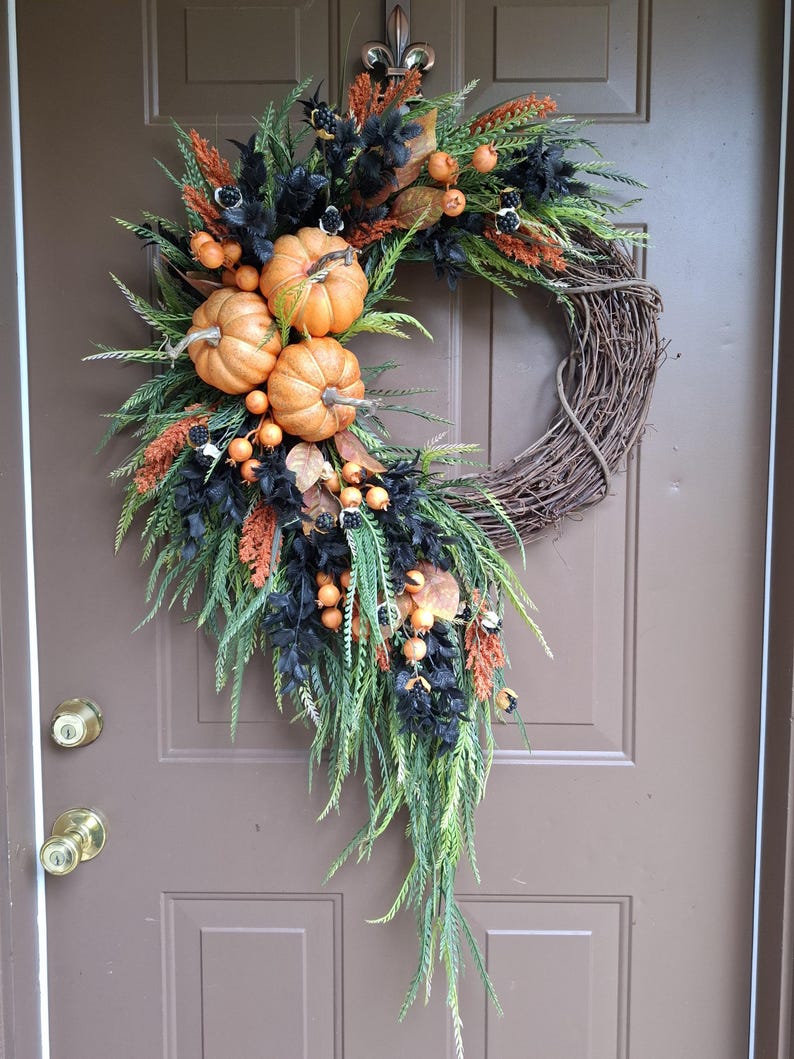 May include: A decorative wreath featuring a circular brown twig base adorned with three orange pumpkins, clusters of small orange berries, black foliage, and green fern-like leaves. The arrangement is asymmetrical, extending downward from the left side.