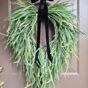 May include: A teardrop-shaped wreath composed of layered green fern-like foliage in varying shades of light and dark green. The wreath is suspended by a long, solid black velvet ribbon tied in a bow at the top, hanging from a metal door hook.