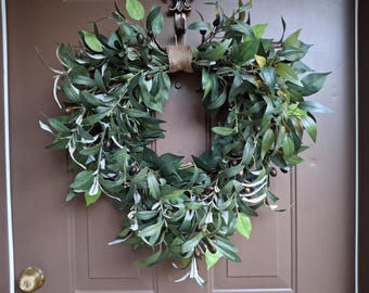 Può includere: Una lussureggiante ghirlanda di ulivo verde con olive scure e foglie verde chiaro, legata con spago, è appesa a una porta marrone. La ghirlanda è un design circolare completo, ideale per l'arredamento della casa.