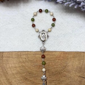 Puede incluir: Un rosario de una decena con un bucle circular de cuentas alternas en rojo, verde y crema conectadas por eslabones de metal plateado. Incluye una medalla ovalada central con una figura, dos peque&ntilde;os dijes de cruz y una cruz celta m&aacute;s grande.