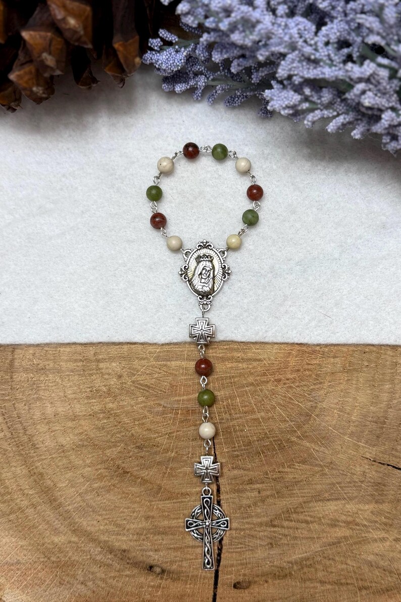 Puede incluir: Un rosario de una decena con un bucle circular de cuentas alternas en rojo, verde y crema conectadas por eslabones de metal plateado. Incluye una medalla ovalada central con una figura, dos peque&ntilde;os dijes de cruz y una cruz celta m&aacute;s grande.