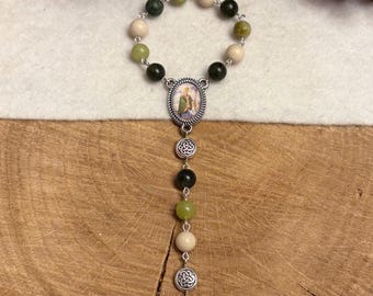 Puede incluir: Un rosario con un bucle circular de cuentas verdes, verde oscuro y beige conectadas por eslabones de metal plateado. Incluye una medalla ovalada central que representa una figura con túnica verde, dos espaciadores celtas plateados y un crucifijo.