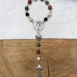 Puede incluir: Un rosario de una d&eacute;cada con una disposici&oacute;n circular de cuentas de piedra multicolor en tonos negro, blanco, verde y marr&oacute;n rojizo. Incluye una medalla de San Benito plateada, dos peque&ntilde;os espaciadores en forma de cruz y un colgante de cruz celta.